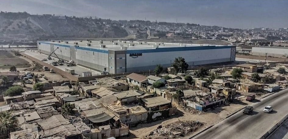 The meme is a split-screen image of two vastly different environments: a sprawling Amazon warehouse on the left, and a densely populated and impoverished neighborhood on the right, with a small sign that reads "Amazon Warehouse" on the right side. The implication is that Amazon's massive warehouse is built on the same land that was previously a community, highlighting the stark contrast between the company's wealth and the poverty of the surrounding neighborhood. The text on the right side is a subtle commentary on the company's priorities and the impact of its business practices.